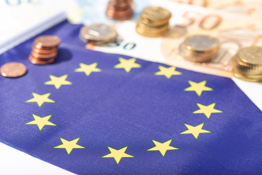 Na stole leży flaga Unii Europejskiej. Obok niej leżą banknoty i monety euro.