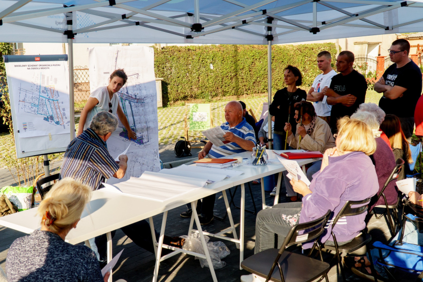 Kilkanaście osób pod dużym namiotem w plenerze. Dyskutują na jakiś temat. Prowadzący dyskusję pokazuje coś na planie osiedla