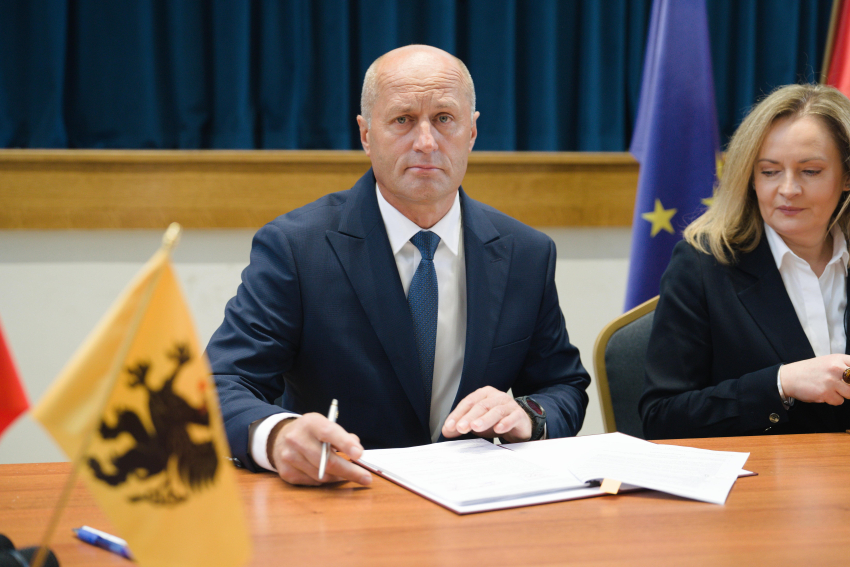 Dwie osoby siedzą przy stole prezydialnym i podpisują umowę