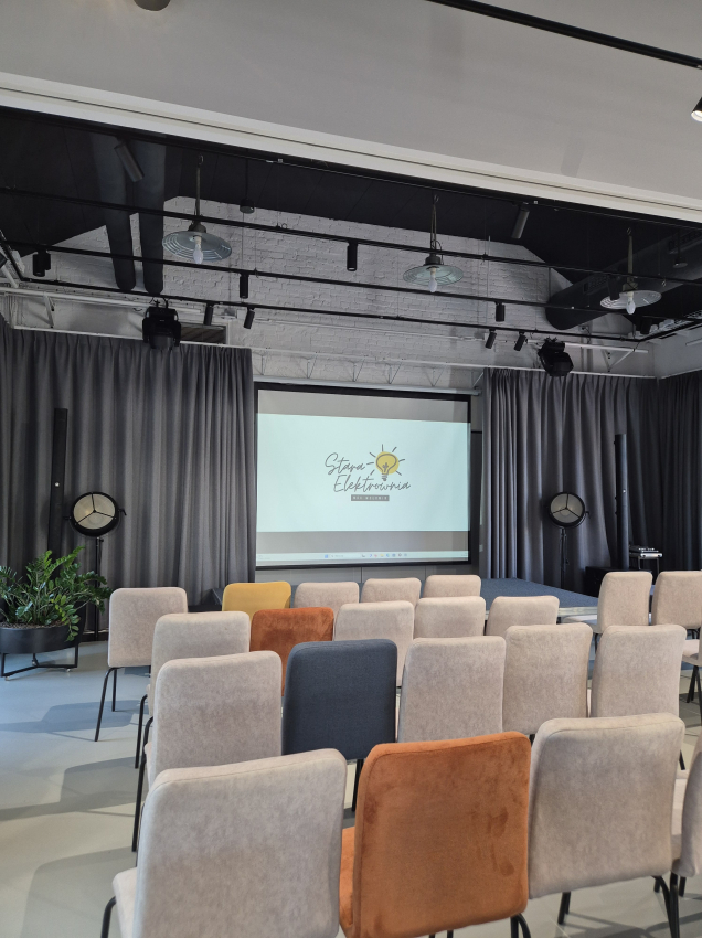 Sala wykładowa wypełniona krzesłami zwróconymi w kierunku pulpitu z prezentacją dotyczącą Starej Elektrowni