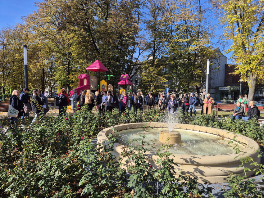 Fontanna wśród krzewów, w tle grupa ludzi, za nimi plac zabaw i drzewa