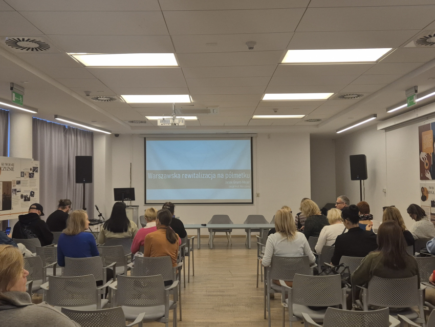 Sala wykładowa wypełniona ludźmi siedzącymi na krzesłach, zwróconymi w kierunku pulpitu z prezentacją dotyczącą rewitalizacji