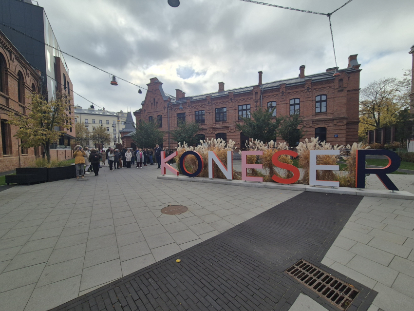 Wyremontowany plac wyłożony betonową kostką. Na środku placu trójwymiarowy napis &quot;KONESER&quot;, w tle grupa ludzi i wyremontowane kamienice z czerwonej cegły