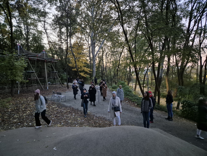 Grupa ludzi chodząca po parku wśród drzew, w tle mały park linowy