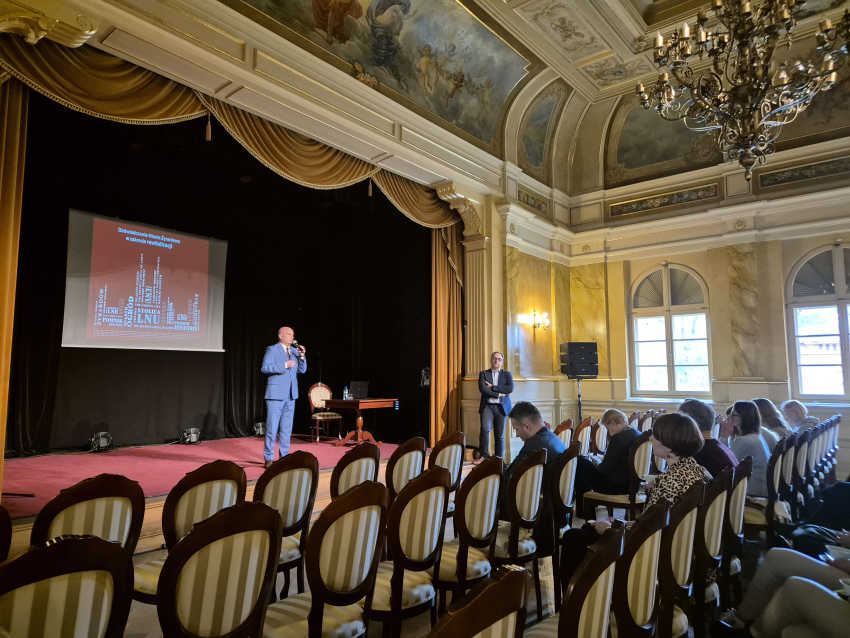Sala teatralna wypełniona krzesłami, na scenie mężczyzna prowadzący prezentację