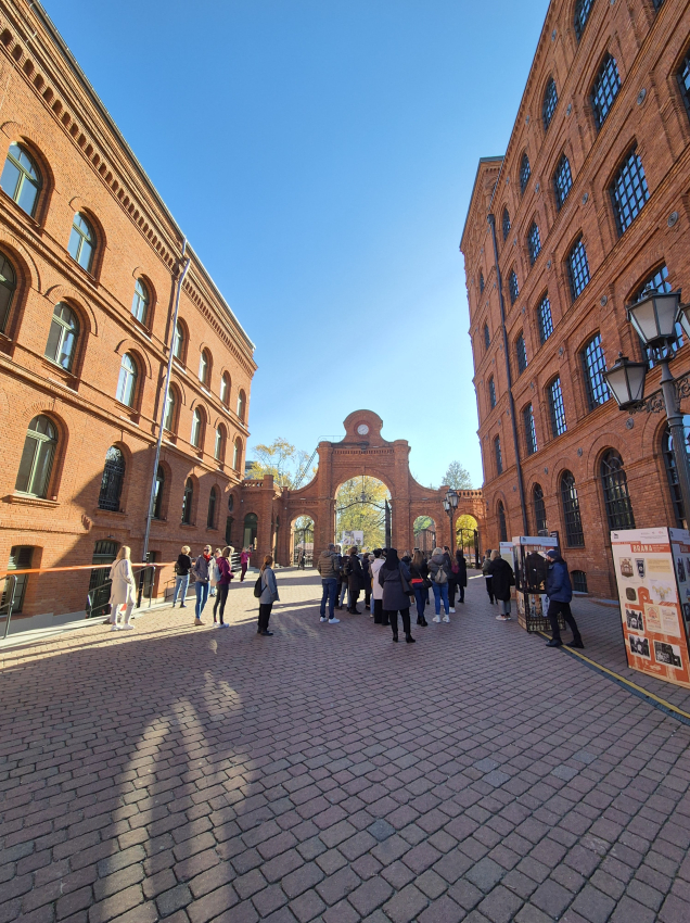 Grupa ludzi na placu pomiędzy budynkami z czerwonej cegły. W tle brama z czerwonej cegły.
