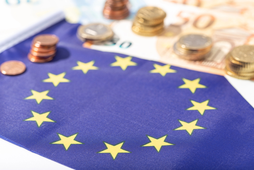 Na stole leży flaga Unii Europejskiej. Obok niej leżą banknoty i monety euro.