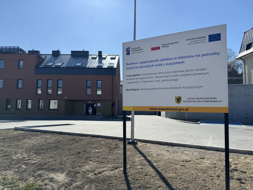 Tablica informująca, że budynek wspólnoty oraz jego wyposażenie zostały dofinansowane z Unii Europejskiej, w tle budynek wspólnoty