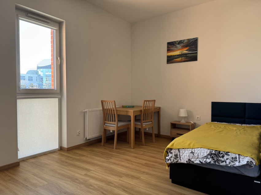 Pokój dla osoby z autyzmem w budynku. W ujęciu łóżko, stół z krzesłami i okno.