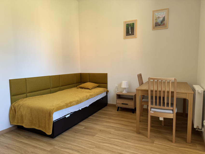Pokój dla osoby z autyzmem w budynku. W ujęciu łóżko, stół z krzesłami na ścianie dwa obrazki