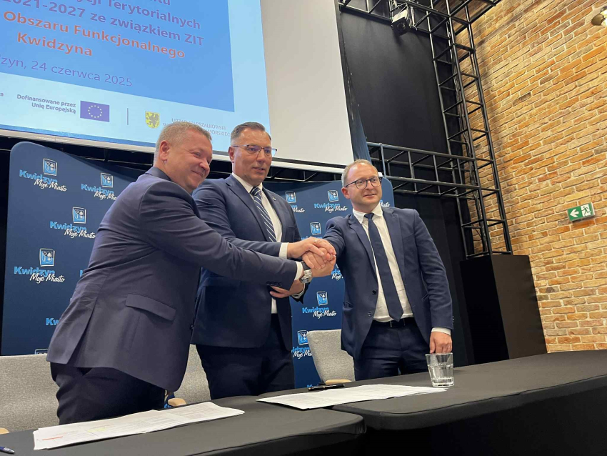 Zdjęcie przedstawia trzech mężczyzn ubranych w garnitury przy stole prezydialnym składających na środku ręce w geście gry drużynowej.