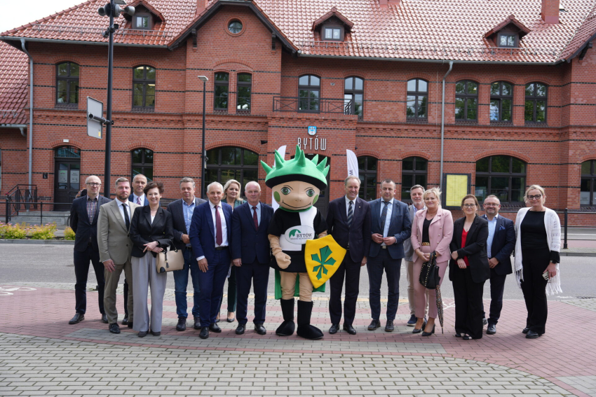 Marszałek Województwa Pomorskiego oraz inni ludzie pozują wraz z człowiekiem ubranym w strój maskotki gminy Bytów. W tle widać dworzec Bytowa.