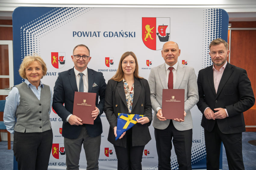 Pięc osób na tle ścianki. Marszałek i startosa trzymają teczkę z umową. Kobieta w środku trzyma flage Unii.