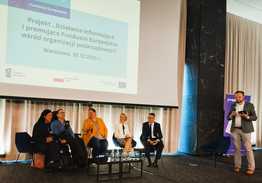 Pięcioro ludzi siedzi na scenie. Po prwej stronie mężczyzna z notatnikiem. W tle ekran.