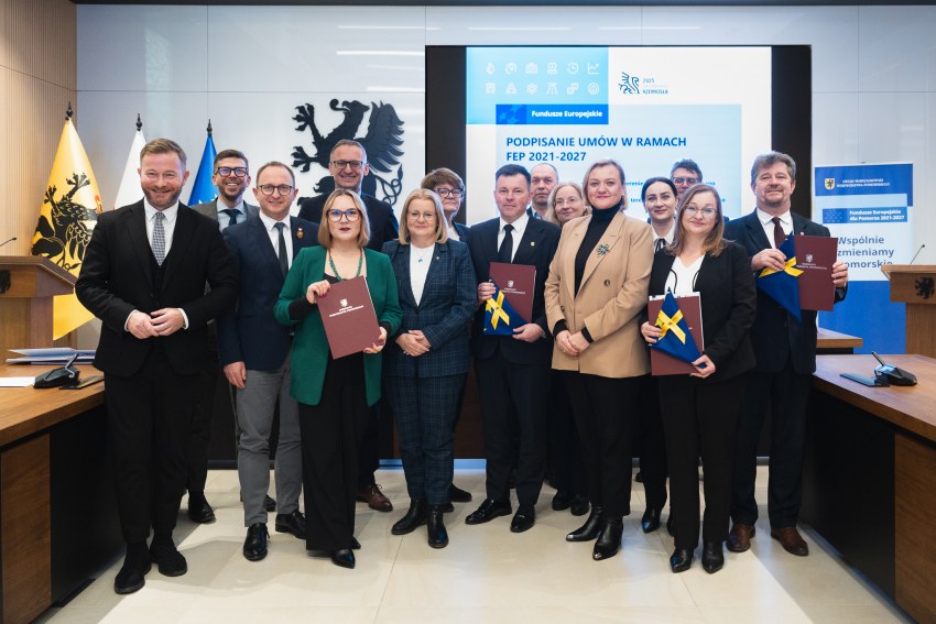 Piętnastu uczestników uroczystego podpisania umów w ramach programu FEP 2021-2027 pozuje do zdjęcia prezentując podpisane umowy oraz flagi unijne