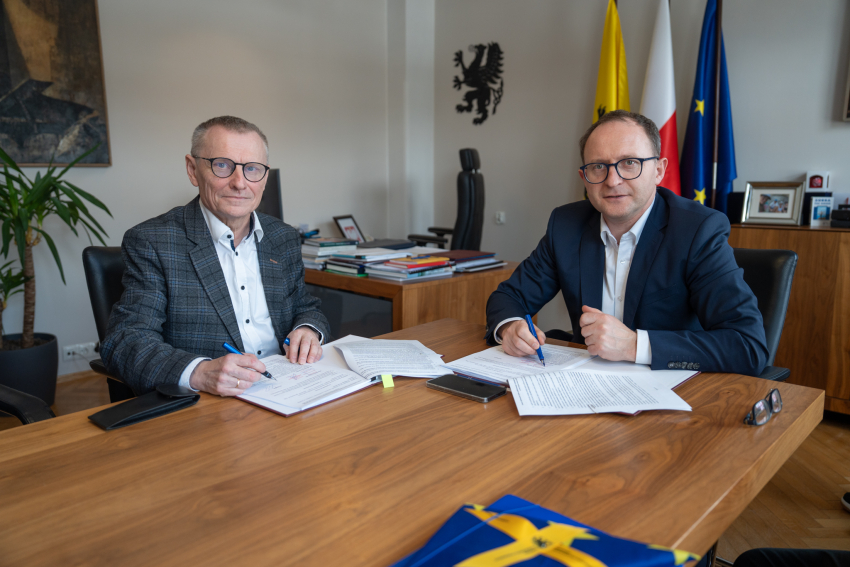 Dwie osoby podczas spotkania przy stole w biurze – dwie siedzą naprzeciw mężczyzny w garniturze, na stole leżą dokumenty, kubki z kawą i telefon.