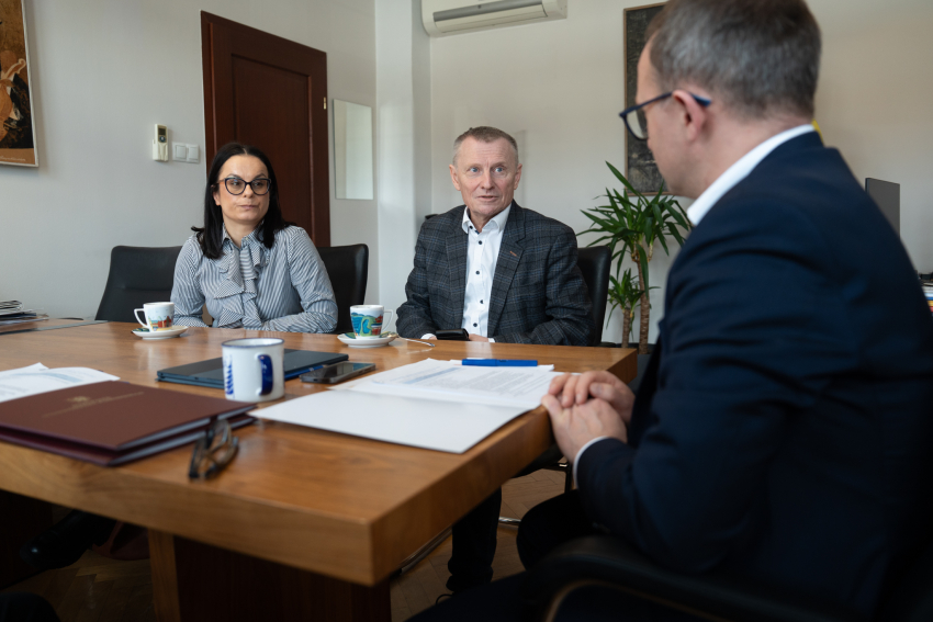 Trzy osoby podczas spotkania przy stole w biurze – dwie siedzą naprzeciw mężczyzny w garniturze, na stole leżą dokumenty, kubki z kawą i telefon.