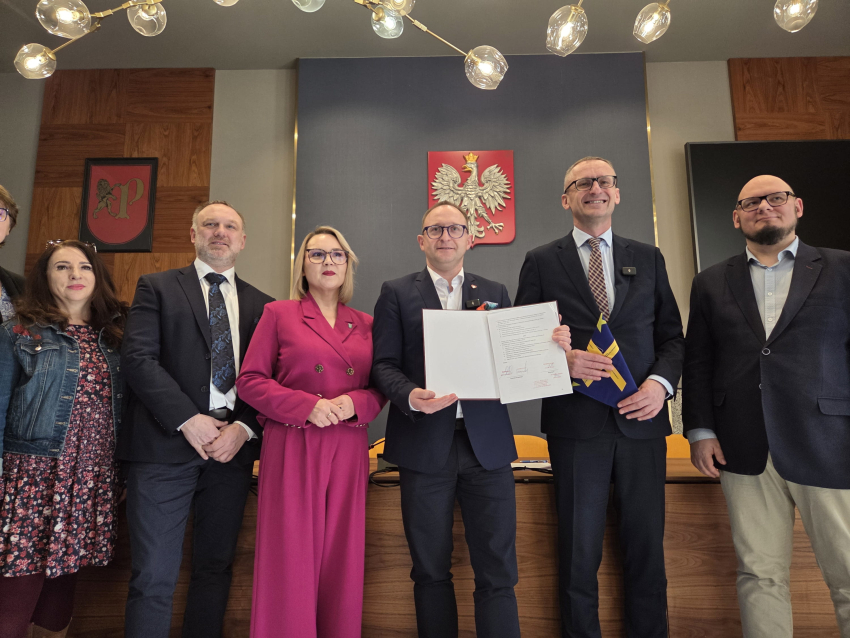 Grupa sześciu osób w strojach wizytowych stoi w rzędzie w oficjalnym pomieszczeniu. Centralna postać, mężczyzna w ciemnym garniturze i okularach, trzyma otwarty, podpisany dokument. Po jego prawej stronie stoi kobieta w wyrazistym, amarantowym garniturze. Na ścianie za nimi widnieje godło Polski (orzeł biały na czerwonej tarczy) oraz herb z lwem. W tle widać nowoczesne oświetlenie sufitowe oraz drewniane panele ścienne. Oficjalne podpisanie porozumienia lub uroczystość urzędową.