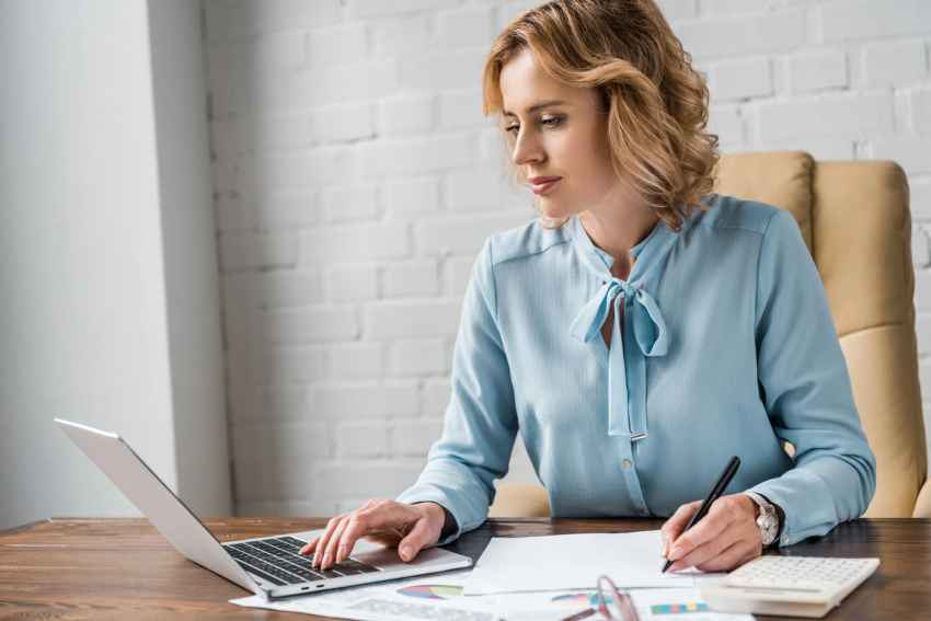 Kobieta trzyma długopis. Drugą ręką pisze coś na klawiaturze laptopa. 