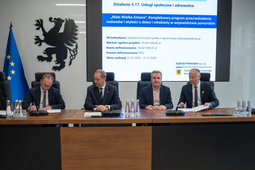 Cztery osoby podpisują umowę przy stole. W tle prezentacja. 