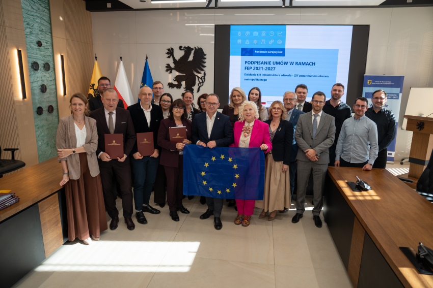 Dwadzieścia osób pozuje do uroczystego zdjęcia, prezentując podpisane umowy oraz rozwiniętą flagę Unii Europejskiej. Wśród zebranych znajduje się wicemarszałek województwa pomorskiego Marcin Skwierawski