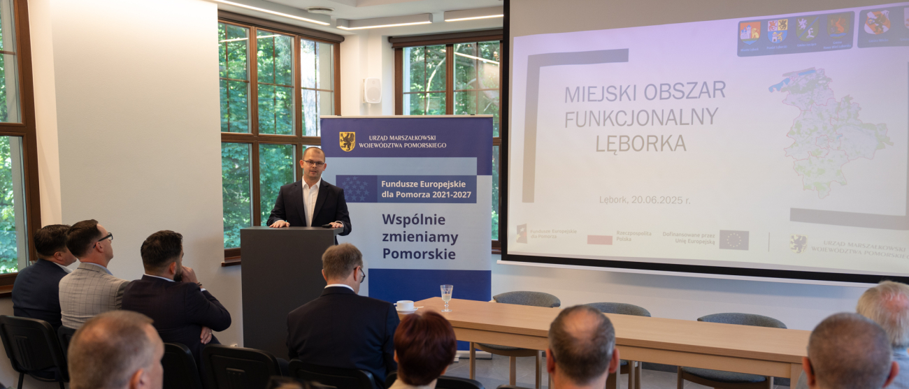 Ktoś stoi przy mównicy i przemawia do zebranych. W tle obraz rzutnika oraz rollup "Wspólnie zmieniamy Pomorskie"