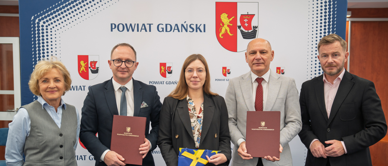 Pięc osób na tle ścianki. Marszałek i startosa trzymają teczkę z umową. Kobieta w środku trzyma flage Unii.