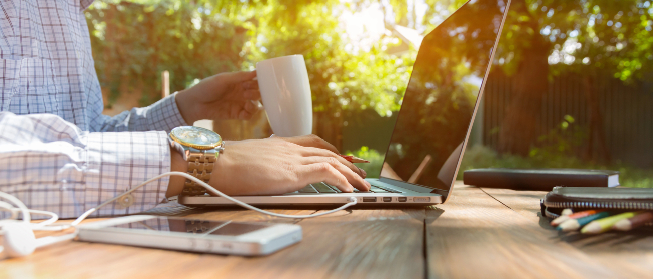 Człowiek przed laptopem. Widać jego rękę. Na stole leży telefon i długopisy