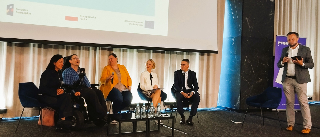 Pięcioro ludzi siedzi na scenie. Po prwej stronie mężczyzna z notatnikiem. W tle ekran.