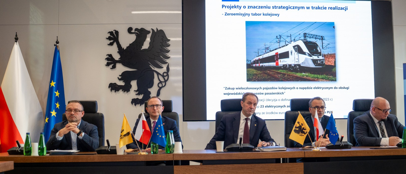 Pięć osób przy stole. W tle flagi i ekran