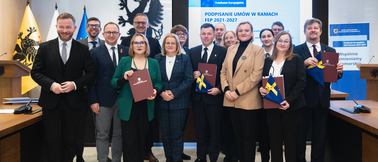 Piętnastu uczestników uroczystego podpisania umów w ramach programu FEP 2021-2027 pozuje do zdjęcia prezentując podpisane umowy oraz flagi unijne