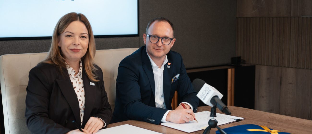 Dwie osoby w strojach biznesowych, kobieta i mężczyzna, siedzą przy drewnianym stole i podpisują dokumenty. Oboje uśmiechają się w stronę obiektywu. W tle widoczny jest duży ekran z napisem „GDYNIA 2026”, a na stole przed nimi stoi mikrofon z logotypem oraz flaga UE