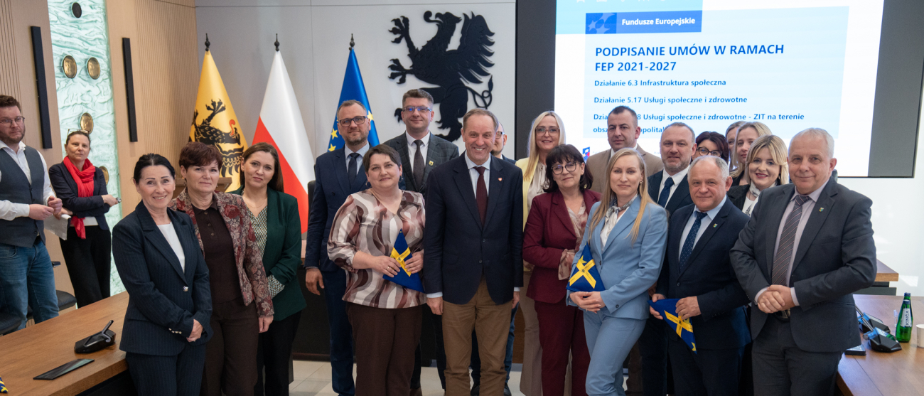 dwadzieścia jeden osób pozuje do zdjęcia prezentując podpisane umowy oraz trzymając flagi unijne. Wśród nich marszałek województwa pomorskiego Mieczysław Struk, oraz przedstawiciele lokalnych władz.