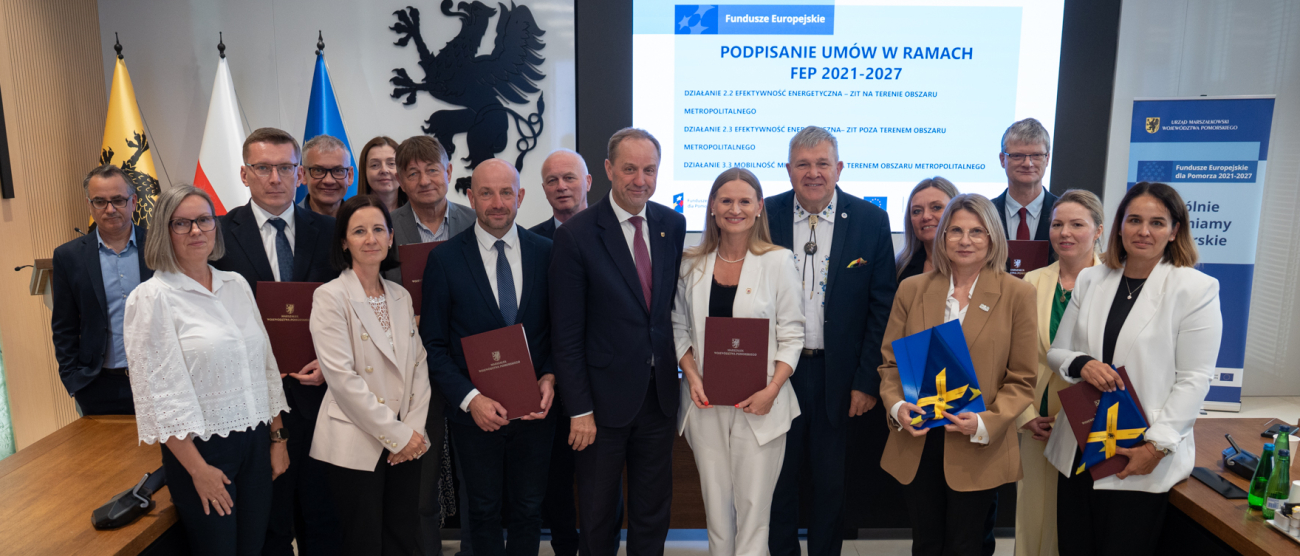siedemnaście osób pozuje do zbiorowego zdjęcia eksponując flagi UE oraz podpisane umowy o dofinansowanie